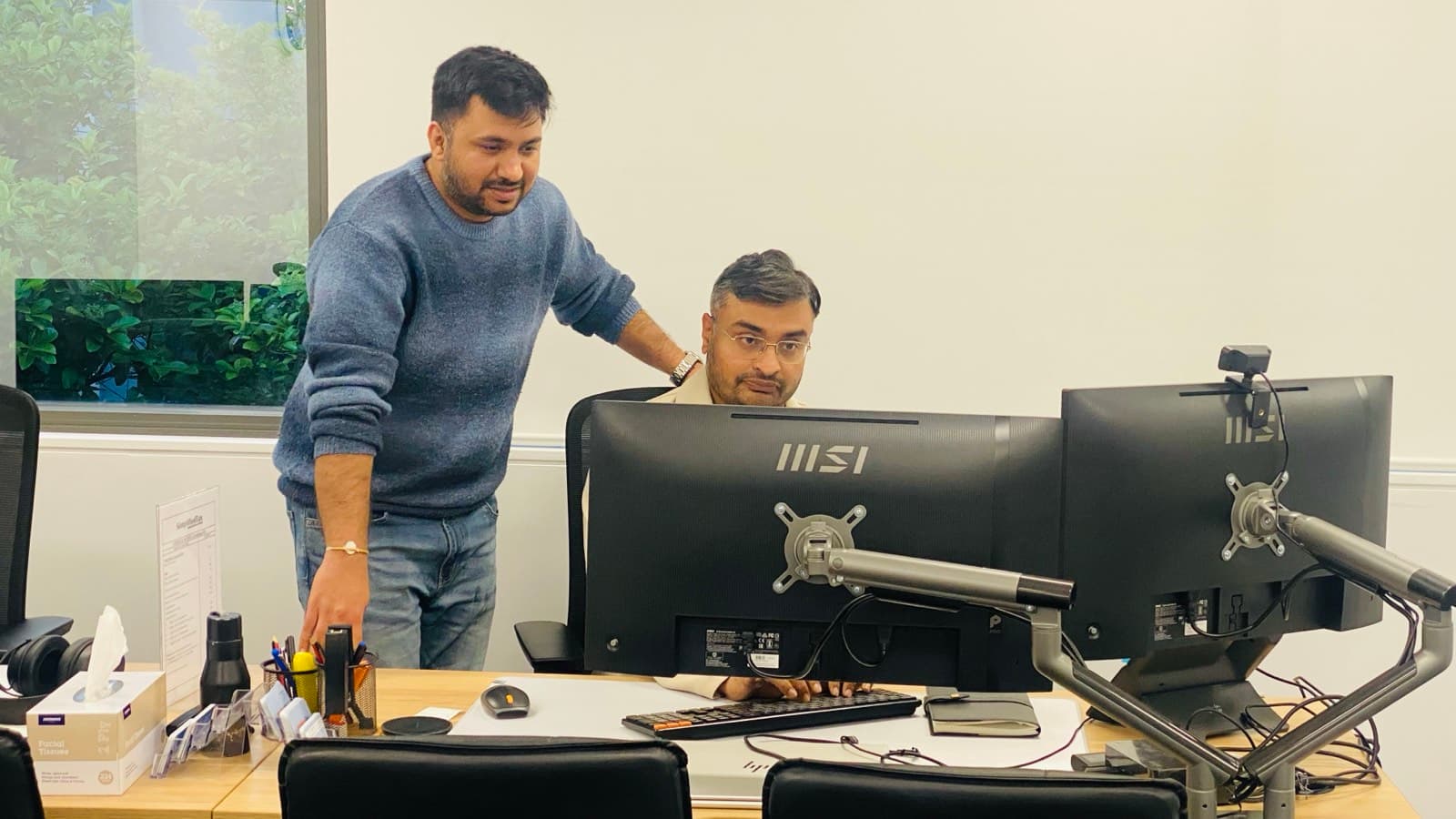 Two people at an office desk with dual monitors, one standing and leaning in to talk through what's on screen with the other.